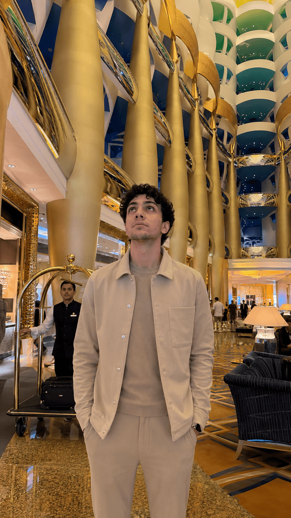 Man in a beige set standing in the gold-columned atrium lobby of the Burj Al Arab, looking up