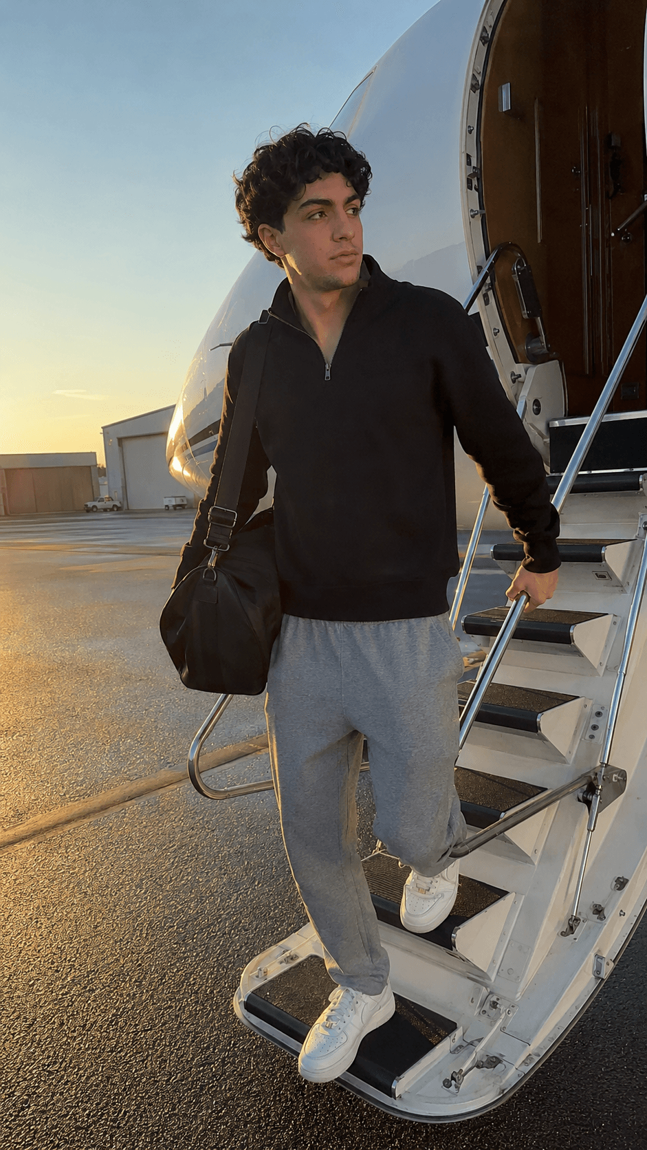 Man in a black quarter-zip and grey sweatpants stepping down the airstair of a private jet at sunset