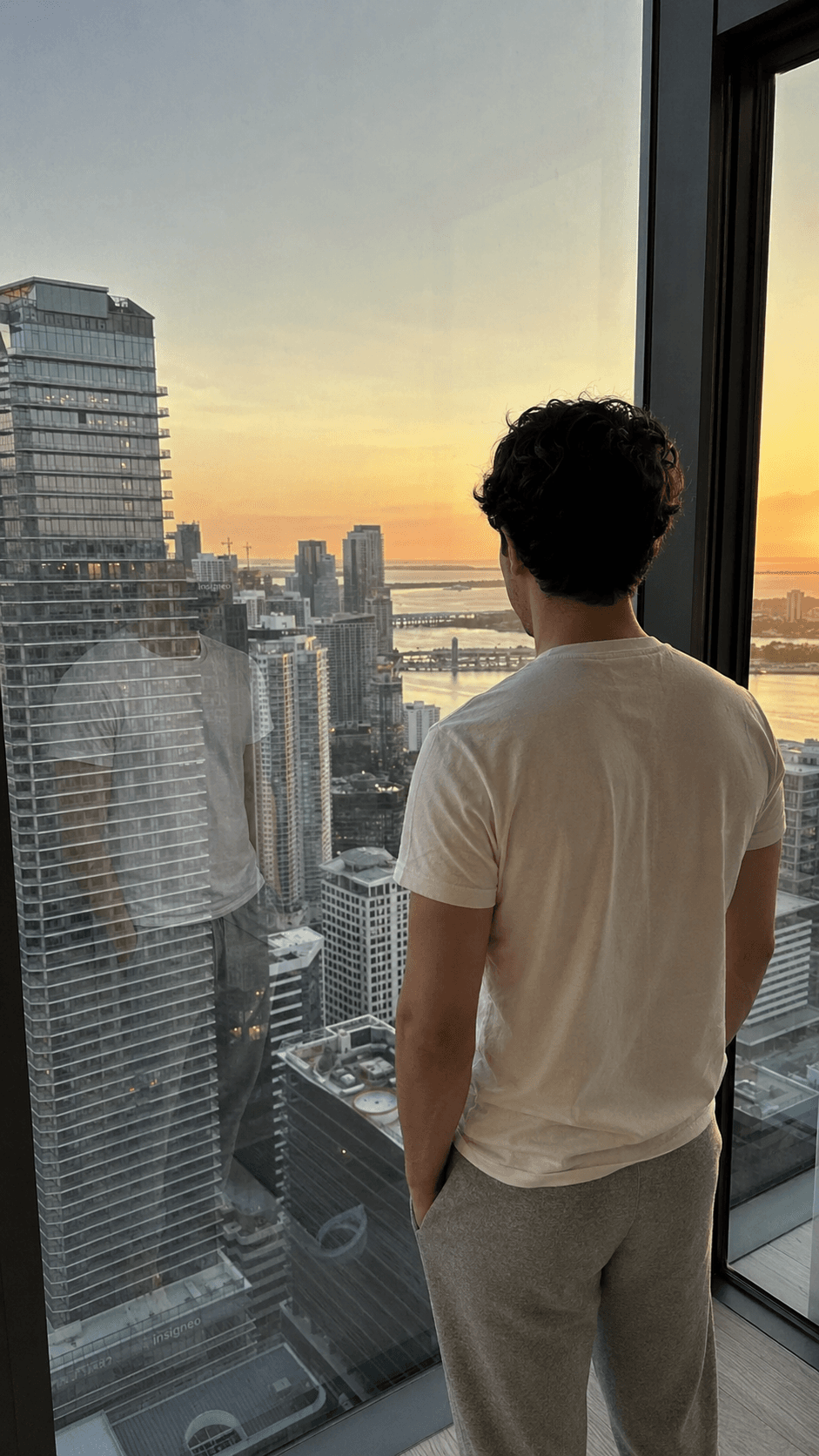 Man seen from behind at a floor-to-ceiling penthouse window overlooking the Miami skyline at sunset