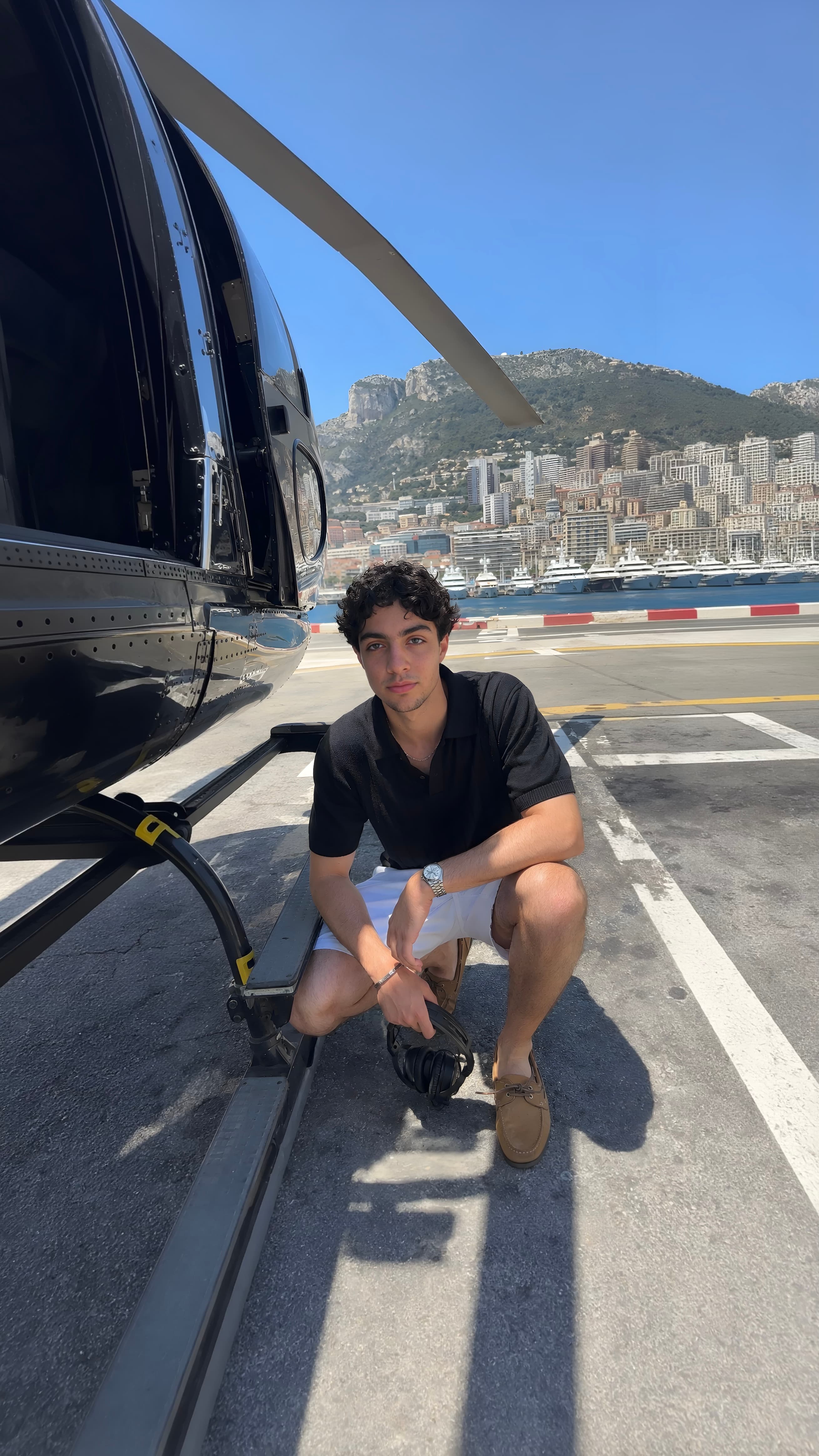 Man crouched beside a black helicopter on the Monaco helipad, the harbour full of superyachts and the city rising behind
